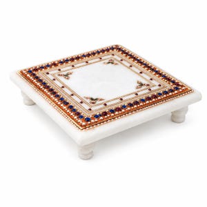 Può includere: Una piattaforma quadrata in marmo bianco con quattro piedini corti e arrotondati. La superficie superiore presenta un bordo complesso con pietre blu e rosse incastonate, dettagli color oro e motivi floreali agli angoli del centro.