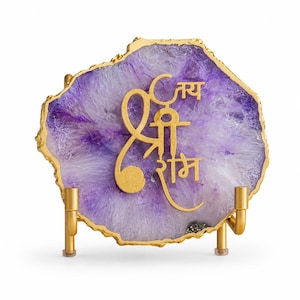 May include: A purple and white agate slice with a gold-coloured metallic edge and a gold-coloured metallic script overlay featuring the Hindi text Jai Shri Ram. The agate slice is supported by a gold-coloured metal stand with two vertical prongs.