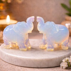 May include: Two opalescent elephant figurines face each other on a marble-like surface. The elephants are carved with detailed features and have a soft, glowing appearance. The background is blurred, with a candle and flowers adding to the serene aesthetic.