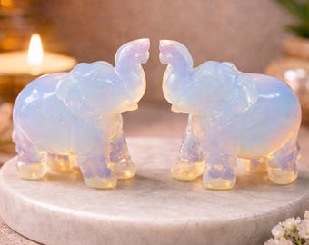 Puede incluir: Dos figuras de elefantes opalescentes se enfrentan sobre una superficie similar al mármol. Los elefantes están tallados con detalles y tienen una apariencia suave y brillante. El fondo está borroso, con una vela y flores.
