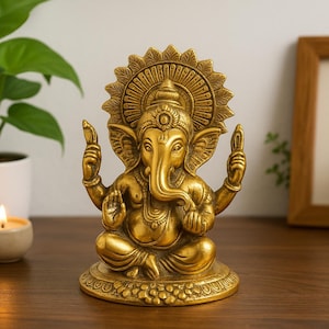 May include: A metallic gold-colored statue of the deity Ganesha, depicted in a seated position with four arms, large ears, and a trunk curved to the left. The figure features an ornate circular halo behind the head and sits on a circular base.