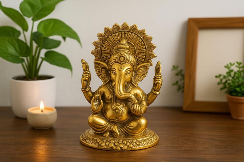 May include: A metallic gold-colored statue of the deity Ganesha, depicted in a seated position with four arms, large ears, and a trunk curved to the left. The figure features an ornate circular halo behind the head and sits on a circular base.