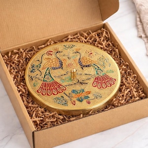 May include: A round, gold-colored decorative box with a lid featuring a peacock design in blue, red, and green. The box is inside a cardboard box filled with brown packing material.