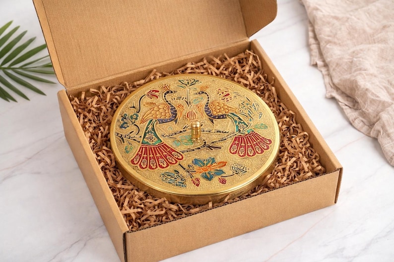 May include: A round, gold-colored decorative box with a lid featuring a peacock design in blue, red, and green. The box is inside a cardboard box filled with brown packing material.