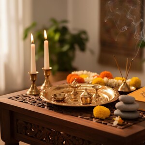 May include: A dark wooden table holds a spiritual display. Two lit candles in brass holders are next to a golden tray with small bowls and a decorative lamp. Incense sticks emit smoke, and a stack of stones and a book are also present.