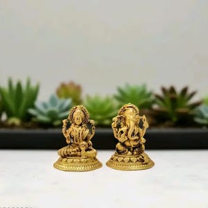 Pode incluir: Duas est&aacute;tuas douradas de divindades hindus. Uma retrata Lakshmi, sentada com v&aacute;rios bra&ccedil;os, e a outra, Ganesha, com cabe&ccedil;a de elefante. Ambas est&atilde;o sobre bases ornamentadas, em um fundo de plantas verdes.