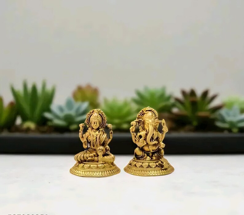 Pode incluir: Duas est&aacute;tuas douradas de divindades hindus. Uma retrata Lakshmi, sentada com v&aacute;rios bra&ccedil;os, e a outra, Ganesha, com cabe&ccedil;a de elefante. Ambas est&atilde;o sobre bases ornamentadas, em um fundo de plantas verdes.