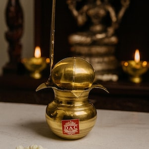 Op de afbeelding: Een gouden messing puja-pot met een deksel en een lange handgreep, met een rode sticker met de tekst "DAC". De pot staat op een wit oppervlak, met een klein trosje witte bloemen ervoor. Twee kleine gouden olielampen en een beeldje staan op de achtergrond.