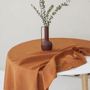 Könnte beinhalten: Ein runder, rostfarbener Tischläufer aus Leinen auf einem weißen Tisch. Eine dunkelbraune Vase mit Henkel enthält grüne Zweige. Der Tisch hat hellfarbene Holzbeine.