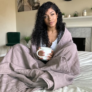 Puede incluir: Una persona sentada en una cama, envuelta en una manta acolchada malva, sosteniendo una taza blanca.