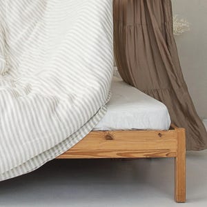 May include: A wooden bed frame with a white fitted sheet and a striped duvet cover in shades of gray and white. A brown fabric canopy hangs on the right side of the bed. The bed is in a room with a white floor.