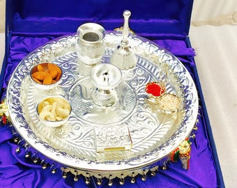 May include: A silver puja thali set with various components, including a small bell, a container, and bowls of nuts. The thali is intricately designed and presented in a blue velvet box, ideal for religious ceremonies.