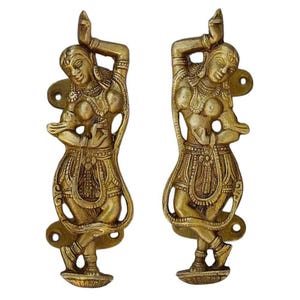 May include: Two antique brass door handles, each featuring a detailed relief of a dancing figure in a traditional pose. The handles are golden-toned, with intricate carvings and mounting hardware.