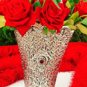Könnte beinhalten: Eine silberne Vase mit einem komplizierten floralen Design und einem roten Juwel in der Mitte. Die Vase ist mit roten Rosen gefüllt.