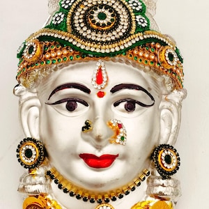 Puede incluir: Una máscara facial decorativa de color plateado con adornos verdes y dorados. La cara está adornada con joyas y un bindi rojo. La máscara está rodeada por una guirnalda de monedas de oro.