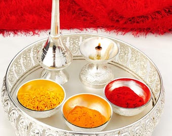 May include: A silver tray with intricate designs, featuring four small bowls filled with yellow, orange, and red powders, a silver diya with a lit wick, and a silver water sprinkler.