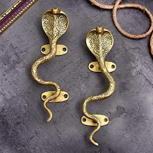 Könnte beinhalten: Zwei goldfarbene Türgriffe in Form von Kobras. Die Griffe weisen aufwendige eingravierte Muster auf und sind auf rechteckigen Platten mit Schraubenlöchern befestigt. Die Schlangenkörper sind geschwungen, und die Kobraköpfe sind erhoben.