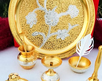 Puede incluir: Una colección de artículos religiosos dorados y plateados, que incluye un plato decorativo grande con diseños florales, una pequeña olla con una cuchara en forma de serpiente, un candelabro, una campana y una figura de vaca. Los artículos están dispuestos sobre una superficie blanca.