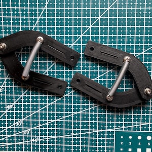 Könnte beinhalten: Zwei schwarze, gebogene, 3D-gedruckte Kunststoffkomponenten, jeweils mit einer silbernen Metallfeder, die durch Schrauben verbunden ist. Die Teile haben eine texturierte Oberfläche mit länglichen Schlitzen und runden Montagelöchern auf einer grünen Rastermatte.