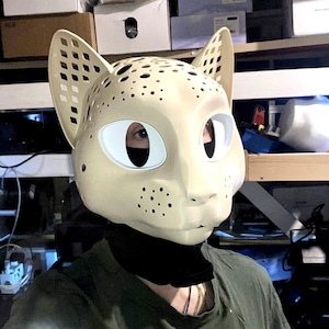 Könnte beinhalten: Eine cremefarbene Katzenmaske mit einem perforierten Gittermuster an den Ohren und kleinen kreisförmigen Aussparungen am Oberkopf. Die Maske hat große weiße Augenöffnungen und eine Reihe kleiner Punkte im Bereich der Schnauze.