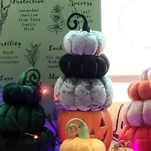 May include: A collection of stacked felt pumpkins in various colours, including green, black, white, orange, and red. The pumpkins are decorated with a swirl design and are arranged on a table with fairy lights.