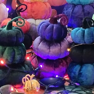 May include: A collection of colourful velvet pumpkins with a gold pumpkin in the foreground. The pumpkins are stacked on top of each other and decorated with fairy lights.