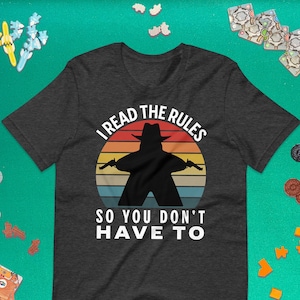 Peut inclure: Un t-shirt gris foncé chiné avec un graphique de coucher de soleil rétro et le texte "I read the rules so you don't have to". Une silhouette d'une personne tenant deux armes à feu est devant le coucher de soleil.