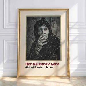 May include: Framed black and white portrait of an elderly person smoking a cigarette. The person is wearing a dark head covering and patterned shawl. The frame is gold, and the text "Her ku mirov here dilê wi li welat dimine" is at the bottom.