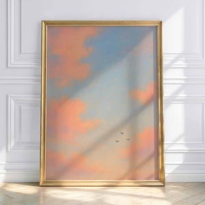 Könnte beinhalten: Ein gerahmter Kunstdruck mit einem pastellfarbenen Himmel mit pfirsichfarbenen Wolken und einem hellblauen Himmel. Drei kleine Vögel fliegen. Das Kunstwerk befindet sich in einem goldenen Rahmen und lehnt an einer weißen Wand.