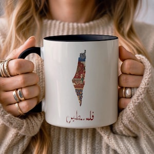 Op de afbeelding: Witte keramische mok met een zwart interieur en handvat. De mok heeft een kleurrijke kaart van Palestina met een decoratief patroon. De tekst "فلسطين" is in het rood op de mok gedrukt.