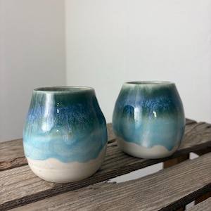 Könnte beinhalten: Zwei handgefertigte, stielose Weingläser aus Keramik. Die Gläser haben eine abgerundete Form mit weißem Boden und einem Farbverlauf aus blau und grün. Die Gläser werden auf einer verwitterten Holzoberfläche präsentiert.