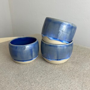 Könnte beinhalten: Drei handgefertigte Keramikschalen. Die Schalen sind glänzend blau mit einem helleren blauen und cremefarbenen Rand. Eine Schale ist separat, während die anderen beiden gestapelt sind. Die Schalen haben eine glatte, abgerundete Form.
