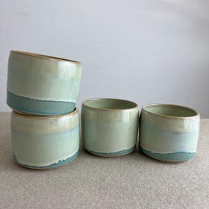 Könnte beinhalten: Vier handgefertigte Keramikbecher mit runder Form. Die Becher sind in Grün- und Blautönen glasiert, mit einem hellbeigen Rand. Die Becher sind gestapelt und auf einer neutralen Oberfläche angeordnet.