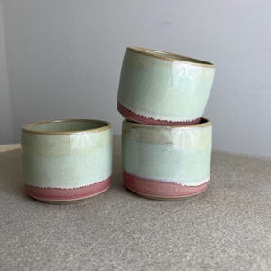 Könnte beinhalten: Drei gestapelte Keramikbecher. Die Becher haben eine zylindrische Form mit einer hellgrünen Glasur auf dem oberen Teil und einem rosa Band am Boden. Die Ränder sind in einem dunkleren Grünton gehalten.