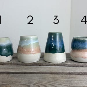 Könnte beinhalten: Vier handgefertigte Keramikvasen mit einzigartigen Farbverläufen. Jede Vase hat einen weißen Sockel, wobei die oberen Abschnitte Farbtöne von Blau, Grün und Rosa aufweisen. Die Vasen sind auf einer Holzoberfläche angeordnet.