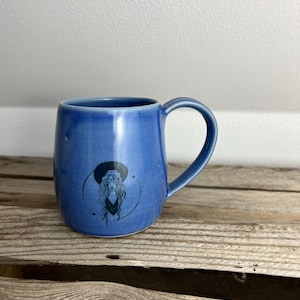 Könnte beinhalten: Eine blaue Keramiktasse mit gebogenem Henkel. Die Tasse zeigt eine schwarze Illustration einer Frau mit langen Haaren und Hut. Die Tasse steht auf einer Holzoberfläche.