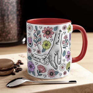 May include: White ceramic mug with a red rim and handle. The mug features a colourful floral design with two hands holding each other. The text "Mummy" and "Olivia" is written on the mug.