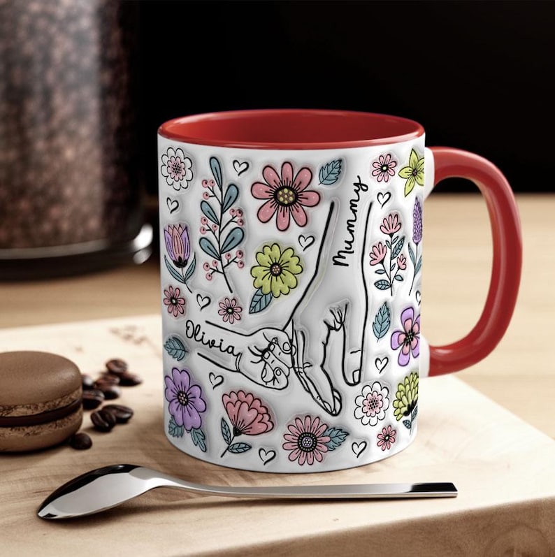 May include: White ceramic mug with a red rim and handle. The mug features a colourful floral design with two hands holding each other. The text "Mummy" and "Olivia" is written on the mug.