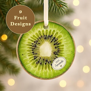Può includere: Un ornamento natalizio a forma di fetta di kiwi con un nastro color crema per appenderlo. L'ornamento ha un esterno verde e marrone con un interno dettagliato. Un piccolo adesivo recita "2025 Amanda". Un cerchio marrone recita "9 Fruit Designs".