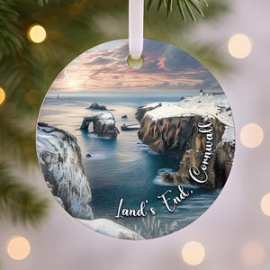 May include: Circular Christmas ornament depicting a coastal scene with cliffs and sea arches. The ornament has a white ribbon for hanging. Text on the ornament reads "Land's End, Cornwall."