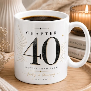 Peut inclure: Un mug en céramique blanche rempli de café noir, portant le texte noir "CHAPTER 40" au-dessus d'un grand chiffre "40", "BETTER THAN EVER", "forty & thriving" en écriture dorée et "EST. 1984" avec des motifs d'étoiles dorées.