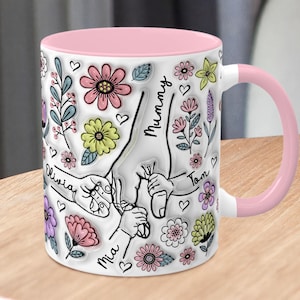 May include: White ceramic mug with a pink rim and handle. The mug features a design of hands holding each other with the names "Mummy", "Tom", "Olivia", "Mia" written on the hands. The design is surrounded by flowers and hearts.