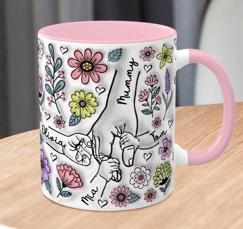 May include: White ceramic mug with a pink rim and handle. The mug features a design of hands holding each other with the names "Mummy", "Tom", "Olivia", "Mia" written on the hands. The design is surrounded by flowers and hearts.