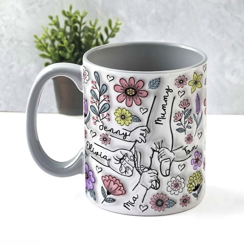 May include: A white ceramic mug with a grey handle. The mug features a floral design with handprints and the names "Mummy", "Denny", "Olivia", "Tom", and "Mia" written on it.