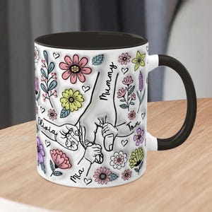 May include: White ceramic mug with a black handle and black rim. The mug features a colourful floral design with handprints and the names "Mummy", "Olivia", "Mia", and "Tom".