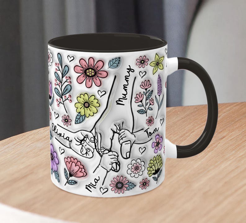 May include: White ceramic mug with a black handle and black rim. The mug features a colourful floral design with handprints and the names "Mummy", "Olivia", "Mia", and "Tom".