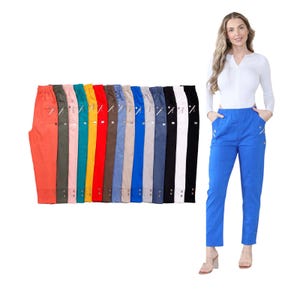 Op de afbeelding: Een verzameling kleurrijke broeken in diverse tinten, waaronder oranje, roze, turquoise en blauw. Elke broek heeft een ritszak en knoopdetails bij de enkel. Een vrouw draagt een blauwe broek met zakken.