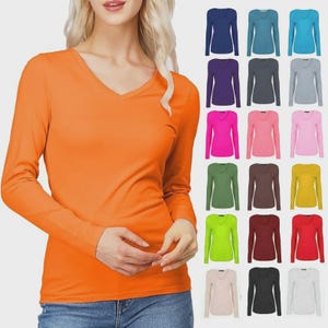 Puede incluir: Una camiseta naranja de manga larga con cuello en V se muestra en una modelo, con una variedad de otras camisetas de manga larga de colores en el fondo. Las camisetas están disponibles en una gama de colores, incluyendo azul, morado, rosa, verde, marrón, amarillo y blanco.