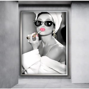 May include: A black and white photo of a woman wearing sunglasses and a white towel on her head. She is applying pink lipstick with a gold applicator.
