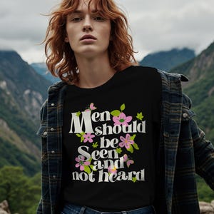 May include: Black t-shirt with the text "Men should be seen and not heard" in white, pink, and green floral design. The shirt is worn by a person with red hair and a denim jacket, set against a mountain backdrop.